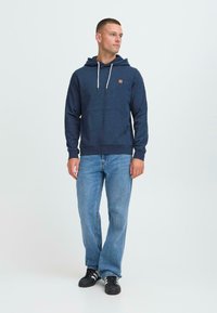 Uomo che indossa un hoodie blu navy, jeans azzurri e sneakers nere con strisce bianche, in piedi e che guarda di lato su uno sfondo bianco.