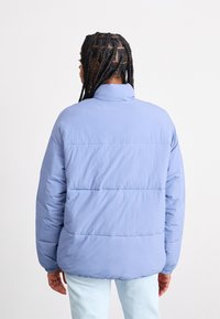 Veste matelassée bleu clair avec un col haut, texture matelassée et poignets élastiques. La vue arrière montre une coupe décontractée et un tissu lisse.
