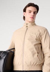 Chaqueta cortavientos beige con cuello alto, cierre de botón doble, bolsillos laterales y dobladillo elástico. Llevada con una mochila negra.