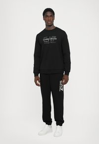 Svart hettegenser og joggebukser laget av en bomullsblanding, med en blank logo på fronten og siden. Hvide sneakers fullfører antrekket.
