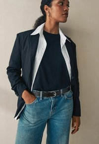 Blazer noir sur une chemise blanche et un t-shirt noir, associé à un jean en denim bleu et une ceinture noire. Tissu lisse et coupe décontractée.