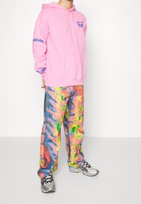 Sudadera rosa con un bolsillo frontal, presenta gráficos azules en el pecho y las mangas. Pantalones anchos de tie-dye multicolor en tonos vibrantes. Zapatillas plateadas.