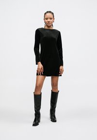 Vestido longo de veludo preto com mangas compridas e decote quadrado, mangas ajustadas, comprimento até a metade da coxa, combinado com botas de couro pretas até ao joelho.