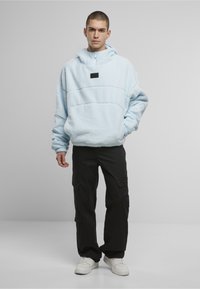 Junger Mann trägt einen hellblauen Fleecepullover mit Tasche vorne, schwarze Cargo-Hosen und weiße Turnschuhe, steht vor einem schlichten Hintergrund.