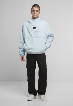 Młody mężczyzna w jasnoniebieskim polarze z kieszenią z przodu, czarnych spodniach cargo i białych trampkach, stojący na jednolitym tle.