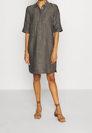 Donna con un vestito di lino grigio carbone al ginocchio con maniche a metà braccio e sandali marroni con tacco e cinturini, in piedi davanti a uno sfondo bianco semplice.