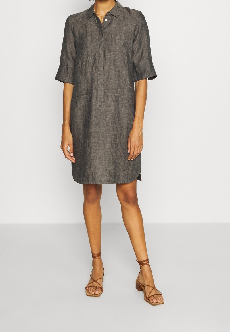 Femme portant une robe en lin gris charbon longueur genoux avec manches mi-longues et sandales à talons marron à brides, debout devant un fond blanc uni.