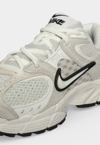 Άσπρο και ανοιχτό γκρι αθλητικό παπούτσι Nike με λεπτομέρειες από δίχτυ και σουέντ, μαύρο-άσπρο λογότυπο Nike swoosh στο πλάι και λευκά κορδόνια.