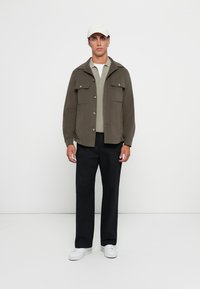 Junger Mann, der eine beigen Mütze, eine olivgrüne Jacke über einem hellgrauen Pullover, schwarze Hose und weiße Sneakers trägt, vor einem weißen Hintergrund steht.
