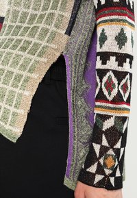 Maglione a maglia con motivi multipli che presenta texture verdi, viola e nere. I design includono motivi geometrici con strisce e colori variati.