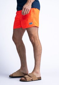 Los bañadores de color naranja brillante presentan un tejido suave y ligero con un parche de logo blanco. Las sandalias de ante marrón con correas ajustables completan el conjunto.