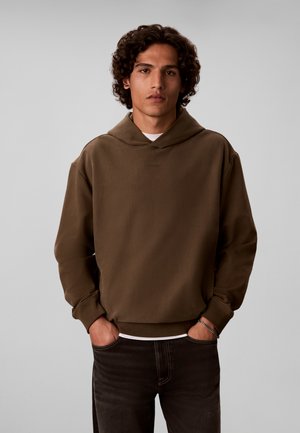 Jeune homme aux cheveux bouclés portant un sweat à capuche marron et un jean foncé, debout les mains dans les poches devant un fond uni.