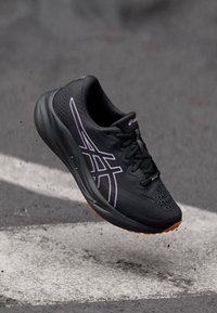 Baskets de course noires avec une tige texturée, présentant un design latéral violet, des lacets noirs et une semelle extérieure en caoutchouc orange. Forme légère et athlétique.