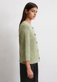 Flauschige grüne Strickjacke mit rundem Ausschnitt, Dreiviertelärmeln und dunklen Knöpfen. Der strukturierte Stoff verleiht Tiefe und Wärme.