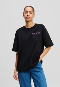 T-shirt en coton noir avec une coupe décontractée, des manches courtes et quatre graphiques de voitures colorés brodés sur le devant. Porté avec un jean en denim bleu.