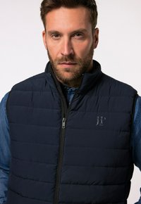 Padded navy Weste mit vertikaler Steppung, hohem Kragen und Frontreißverschluss. Mit Logo auf der linken Brust auf einem glatten Stoffhintergrund.