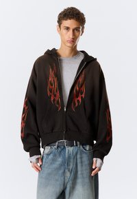 Sweat à capuche noir zippé avec des motifs de flammes rouges sur les manches, porté sur un t-shirt gris. Comprend des poignets côtelés et une capuche avec cordon de serrage.