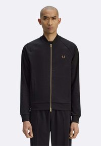 Giacca nera con zip realizzata in tessuto liscio, con collo a coste, tasche laterali e logo dorato sul petto. Vestibilità classica con maniche raglan.