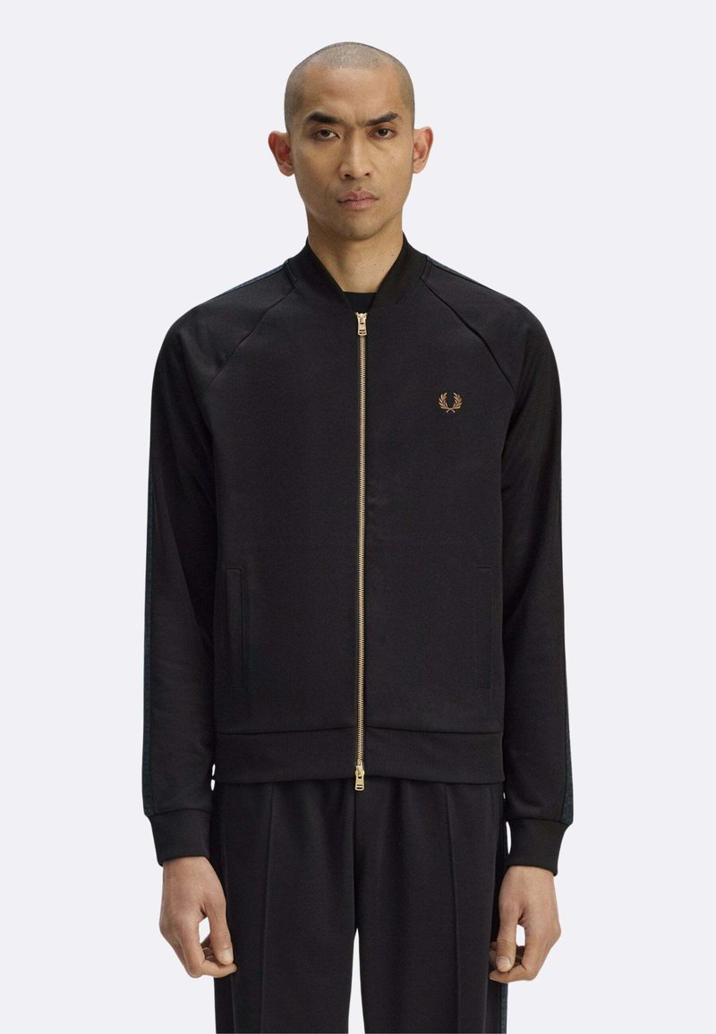 Giacca nera con zip realizzata in tessuto liscio, con collo a coste, tasche laterali e logo dorato sul petto. Vestibilità classica con maniche raglan.