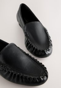 Paire de mocassins loafers en cuir noir avec bordure tressée et bouts arrondis sur une surface claire.
