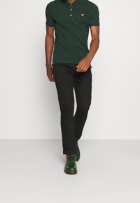 Dunkelgrünes Polohemd mit Knopfleiste, kombiniert mit schwarzer Slim-Fit-Hose und grünen Lederschuhen. Strukturierter Stoff und dezentes Branding.