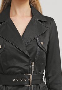 Trench-coat noir avec col à revers, fermeture éclair et taille ceinturée. Présente des garnitures brillantes et une finition lisse et texturée.