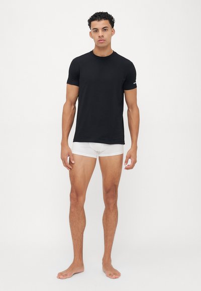 Schwarzes kurzärmliges T-Shirt aus Baumwolle im figurschmeichelnden Stil. Kombiniert mit weißen Boxershorts. Das Model steht barfuß vor einem schlichten Hintergrund.