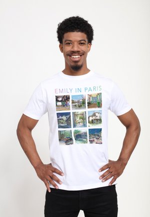 Smilende mand med krøllet hår iført hvid "Emily in Paris" T-shirt med ni farverige Paris-tema fotoruder.