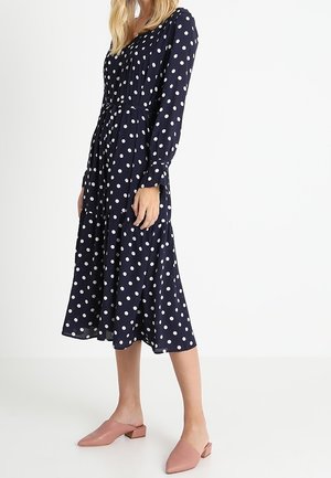 Femme portant une robe midi à manches longues bleu marine à pois blancs et des chaussures à enfiler roses à petit talon, debout sur un sol blanc.