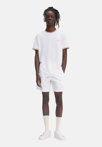 Camisa de tirantes corta blanca con cuello y mangas de canalé multicolor, combinada con pantalones cortos blancos y zapatillas blancas, que presenta un logo en el pecho.
