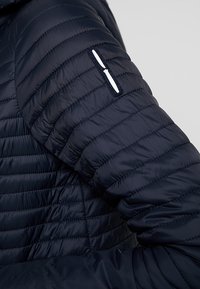 Manga de chaqueta acolchada azul marino con diseño de acolchado horizontal, que presenta un pequeño bolsillo con cremallera y un acento blanco. Textura ligera y suave.