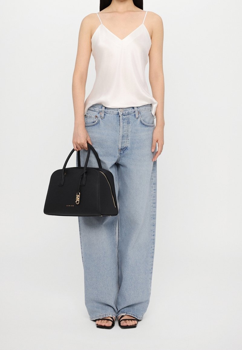 Femme portant une camisole en satin blanc, un jean bleu clair à jambes larges, des sandales noires, tenant un sac à main structuré noir avec un bijou doré.