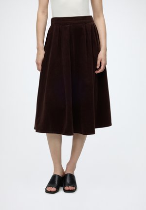 Femme portant une jupe midi plissée marron foncé, un haut blanc et des mules à talons ouvertes noires, debout devant un fond uni.