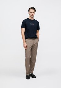 Μπλε βαμβακερό t-shirt με γραφικό λογότυπο, συνδυασμένο με ανοιχτό καφέ παντελόνια και μαύρα παπούτσια. Το σύνολο συνδυάζει casual και επίσημα στοιχεία.