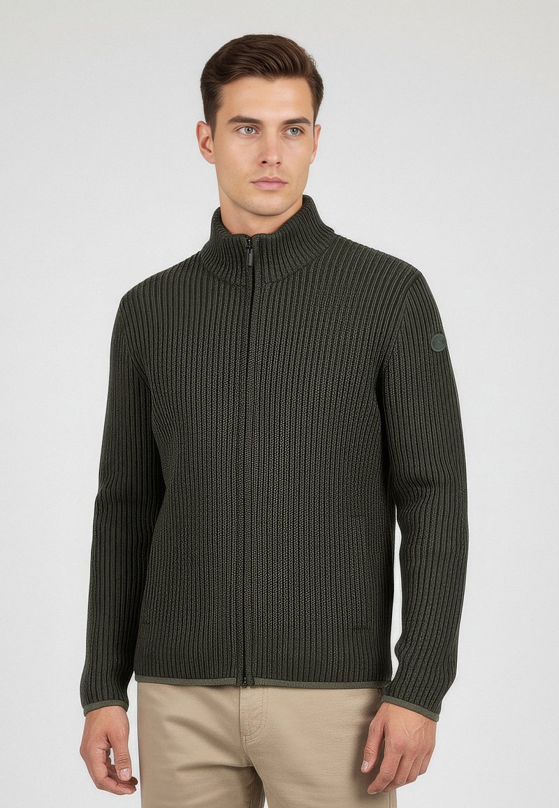 Maglione zip-up verde scuro con texture a costine, alto colletto e una sottile toppa con logo sulla manica sinistra. Indossato sopra pantaloni beige.