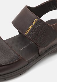 Brun lädersandal med justerbara remmar, texturerad yta och präglad logotyp. Har en vadderad innersula och gummisula för komfort.