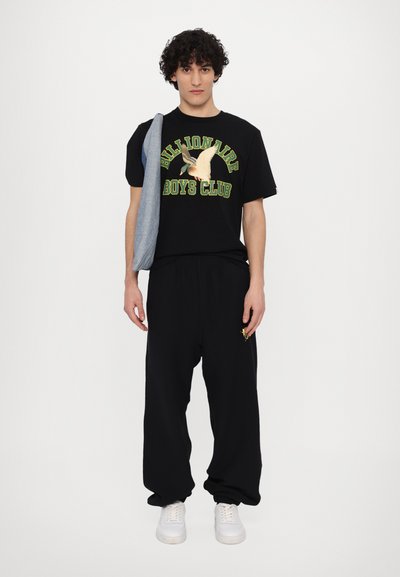T-shirt en coton noir avec un graphique coloré "Billionaire Boys Club" et un pantalon de survêtement noir à coupe décontractée. Le modèle tient une veste en denim. Baskets blanches.