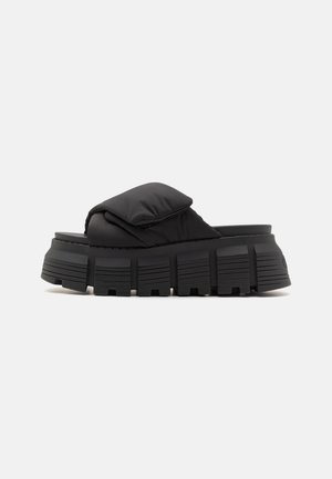 Chancla negra con parte superior acolchada y suela de goma gruesa y estriada. Presenta un diseño minimalista y una forma redondeada para el soporte del pie.