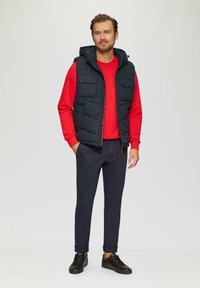 Marineblaue Steppweste mit Kapuze, ausgestattet mit zwei Brusttaschen, über einem roten Langarmshirt getragen, kombiniert mit dunklen Hosen und schwarzen Schuhen.