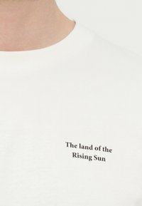 Λευκό μπλουζάκι με στρογγυλή λαιμόκοψη που δείχνει το κείμενο "Η χώρα του Ανατέλλοντος Ηλίου" με μικρά μαύρα γράμματα στο αριστερό μέρος του στήθους.