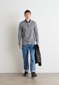 Camisa pólo cinza de mangas longas com colarinho e pala de três botões, combinada com jeans azul claro e sapatos pretos, segurando uma jaqueta preta.