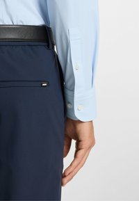 Camicia a righe blu chiaro con bottoni bianchi, abbinata a pantaloni navy con una cintura nera e una piccola etichetta con logo sulla tasca.