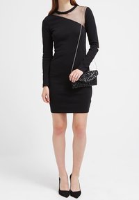 Robe noire cintrée avec manches longues et détail en tulle transparent au niveau du décolleté. Associée à un sac à main noir à sequins et des talons hauts.