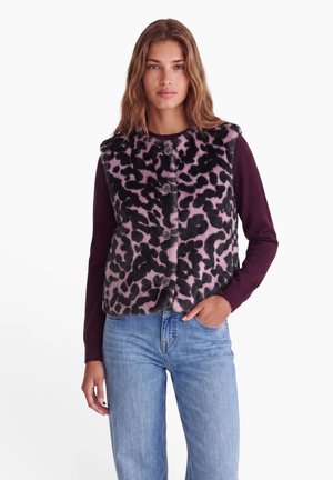 Donna che indossa un gilet viola con stampa leopardata abbottonato sopra una maglia a maniche lunghe viola scuro e jeans azzurri chiari, in piedi con una mano in tasca.