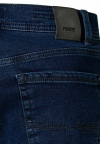 Granatowe dżinsy z denimu z skórzaną łatą oznaczoną napisem "Pioneer" na pasku w talii oraz widocznymi detalami przeszycia tylnej kieszeni.