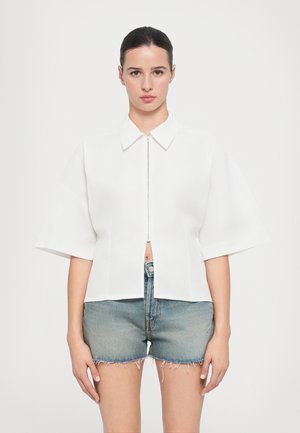 White short-sleeve blouse with a collar and front zipper, featuring a cropped design. Paired with distressed denim shorts.