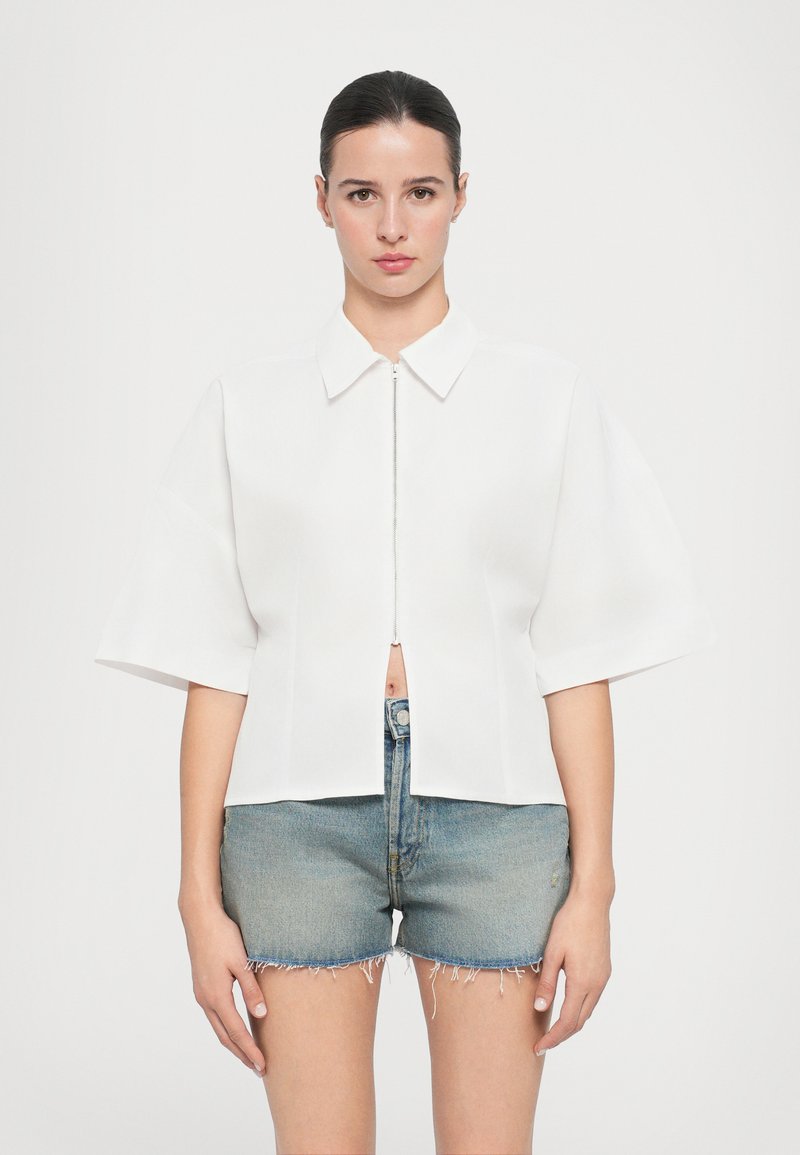 Witte blouse met korte mouwen, een kraag en een rits aan de voorkant, met een crop ontwerp. Gepaard met versleten denim shorts.