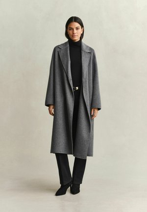 Frau steht und trägt einen langen grauen Mantel über einem schwarzen Rollkragenpullover, schwarze Hose und schwarze Absatzschuhe vor einem schlichten beigen Hintergrund.