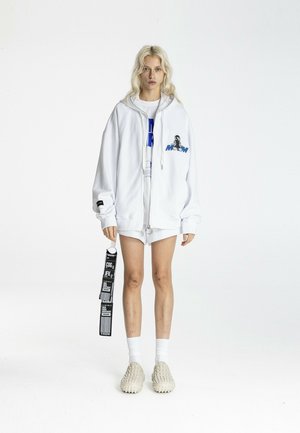 Persona rubia de pie con sudadera blanca con cremallera grande con gráfico, pantalones cortos blancos, calcetines y zapatos slip-on beige texturizados, sosteniendo una etiqueta negra.