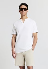 Camicetta henley bianca a maniche corte con due bottoni marroni al colletto, abbinata a pantaloni corti beige texturizzati. Modello con occhiali da sole scuri.